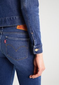 Sininen farkkutakki, jossa on kapea muotoilu, napitettavat hihansuut, takatasku sydämen muotoisella ompelulla ja kontrastinen vaalea nahkaleima.