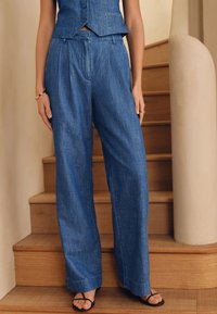 Laisvos, aukšto liemens indigo spalvos džinsinės kelnės su švelnia tekstūra, šoninėmis klostėmis ir tiesiomis kojų linijomis, derinamos su paprastomis juodomis sandalais.