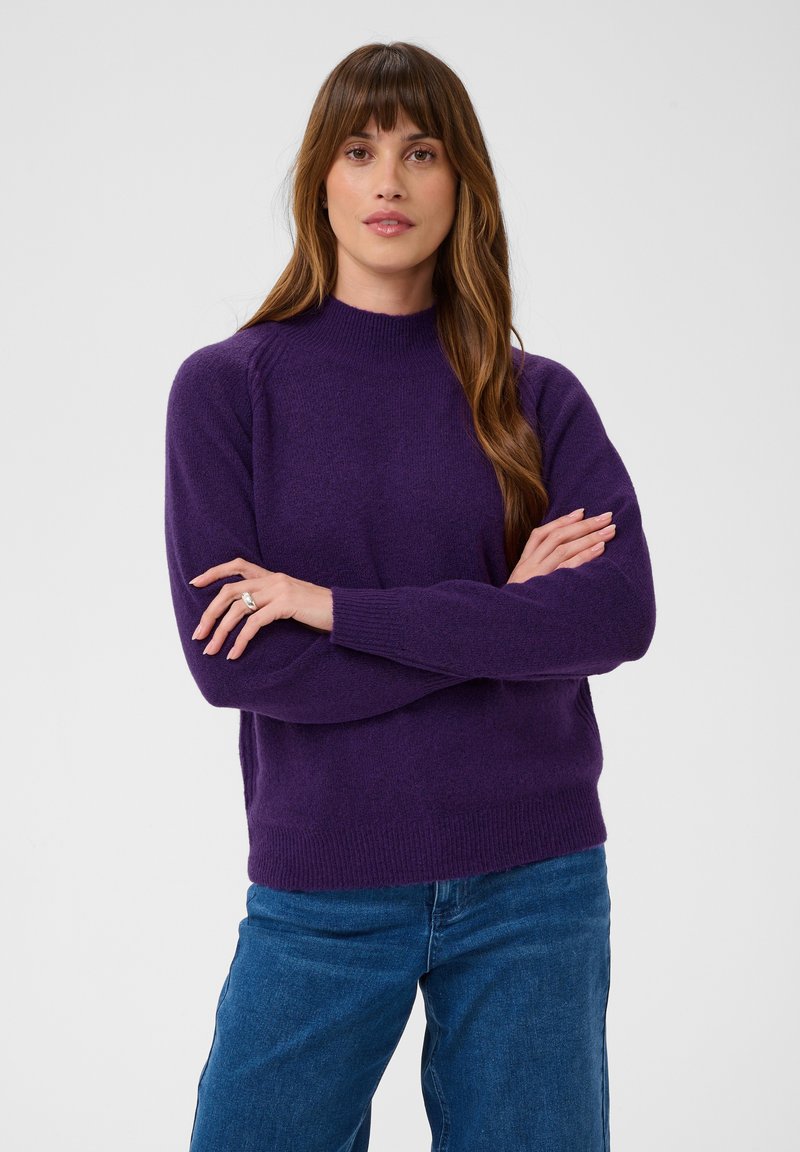 Femme aux longs cheveux bruns portant un pull violet et un jean bleu, debout les bras croisés devant un fond clair uni.