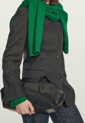 Person trägt ein dunkelgraues Sakko und einen leuchtend grünen Schal, hält eine schwarze Lederhandtasche in der linken Hand.