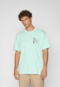 Mintgrøn bomulds t-shirt med rund halsudskæring, korte ærmer og et grafisk design på forsiden, der viser en figur og en stiliseret accent.