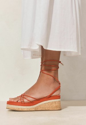 Picior purtând o sandaletă cu platformă și barete portocalii, talpă de plută, cu legături în jurul gleznei și tivul unei fuste albe fluide deasupra gleznei.