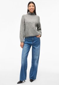 Suéter cinza em padrão espinha de peixe com decote alto, combinado com jeans azul de corte largo e sapatos de salto stiletto pretos.