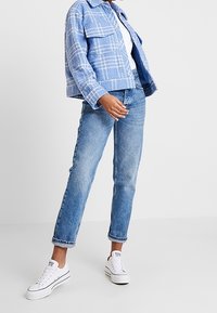 Blå rutig kortjacka över en vit skjorta, kombinerat med ljusblå raka jeans och vita sneakers. Outfiten kännetecknas av avslappnad och ledig stil.