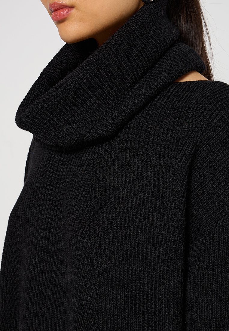 Frau trägt einen schwarzen, dick gestrickten Pullover mit einem großen, umgeschlagenen Rollkragen, der von der Schulter bis zum Kinn sichtbar ist.