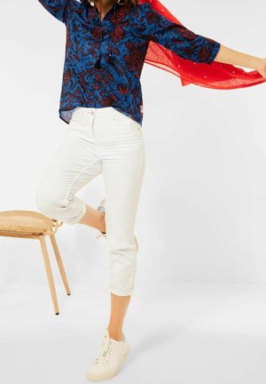 Chemise boutonnée à fleurs bleues avec des accents rouges, associée à un jean skinny blanc retroussé aux chevilles et des baskets de couleur claire.