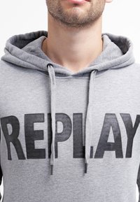 Felpa grigia realizzata in tessuto morbido, caratterizzata da grandi scritte nere "REPLAY" sul davanti. Include un cappuccio con coulisse e una consistenza sottile.