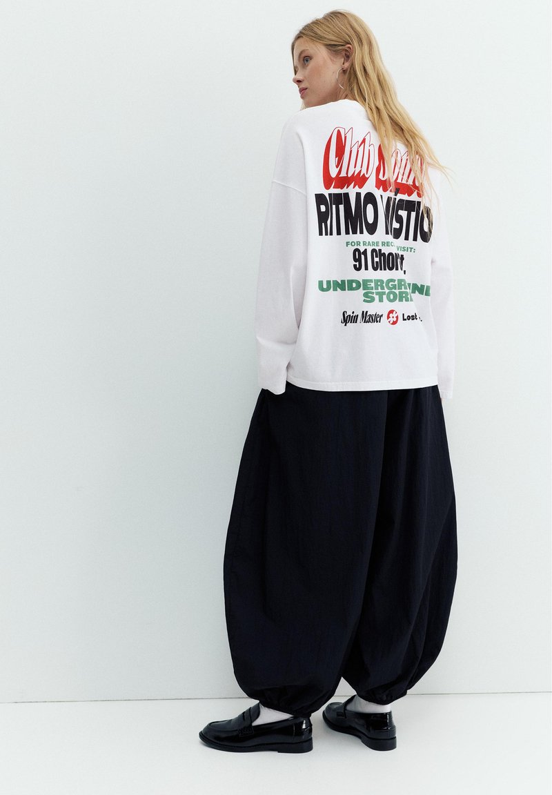 Femme aux longs cheveux blonds portant un t-shirt à manches longues blanc avec un motif graphique et un pantalon noir ample, debout contre un mur blanc uni.