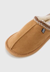 Chausson en suède marron à enfiler avec des détails cousus et un intérieur en fausse fourrure crème douce sur fond blanc.