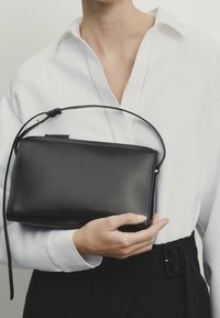 Borsa a tracolla in pelle nera con una superficie liscia, forma rettangolare, manico corto e chiusura con zip, tenuta da una persona in camicia bianca.