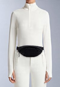 Svart vadderad midjeväska med en elegant design, med en slät yta och justerbar rem, bärs över en vit långärmad topp och byxor.