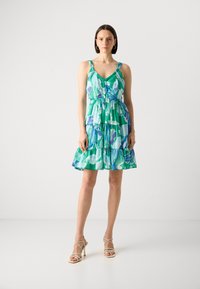 Robe à motifs floraux verts et bleus avec des bretelles spaghetti, taille froncée et jupe à volants ; fabriquée en tissu léger. Sandales beiges.