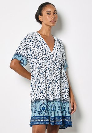 Kobieta w białej sukience z niebieskimi kwiatowymi i ornamentalnymi wzorami, stojąca z jedną ręką na biodrze na tle jednolitym.