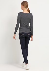 Grijze geribbelde lange mouwtop met gekartelde randen, gecombineerd met donkerblauwe skinny jeans en witte sneakers. Zicht van achteren.