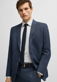 Marineblauw pak met een gestructureerd patroon, gecombineerd met een witte shirt met een klein geometrisch motief en een donkere stropdas. Zwarte riem zichtbaar.