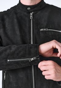 Sort ruskindsjakke med høj krave, der har to lynlåslommer og lynlåse detaljer på ærmerne og lukningen foran. Tekstureret overflade.