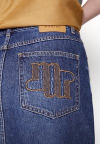 Poche arrière d'un jean en denim avec des lettres stylisées brodées en marron et une étiquette en cuir beige avec le nom de la marque Munthe au-dessus de la ceinture.
