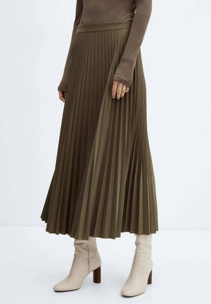 Femme portant une jupe mi-longue plissée vert olive, un haut marron à manches longues et des bottines crème à talons sur fond blanc.