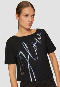 Schwarzes kurzärmliges T-Shirt mit silbernem und blauem Pailletten-Schriftzug. Rundhalsausschnitt, weicher Stoff, figurschmeichelnde Form, minimalistischer Stil.