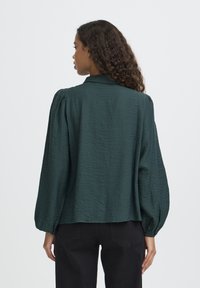 Blusa verde oscuro texturizada con mangas abullonadas, cuello con solapa y corte relajado, combinada con pantalones negros, vista desde atrás.
