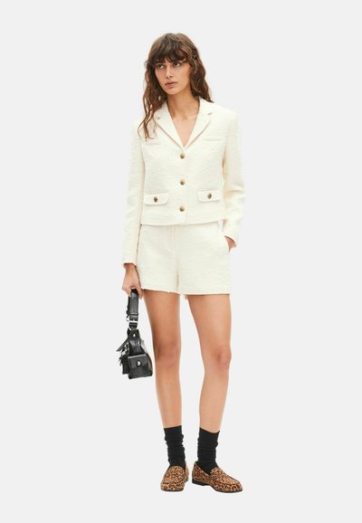 Blazer blanc texturé avec deux poches devant et boutons dorés, associé à un short blanc. Sac à main noir, chaussettes noires et chaussures à imprimé léopard.