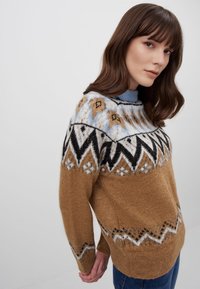 Maglione lavorato a maglia marrone con uno scollo a barca blu, avorio e nero. Presenta design geometrici e un collo rotondo. Tessuto morbido e testurizzato.