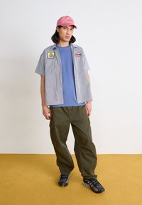 Camicetta a righe con maniche corte sopra una maglietta blu, pantaloni cargo color oliva e sneakers nere. Completata con un cappellino rosa. Visibili toppe sulla camicia.