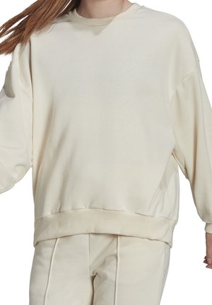 Vrouw met oversized, crèmekleurige sweatshirt en bijpassende broek met geribbelde boorden en zoom, haar dat naar de zijkant wappert.