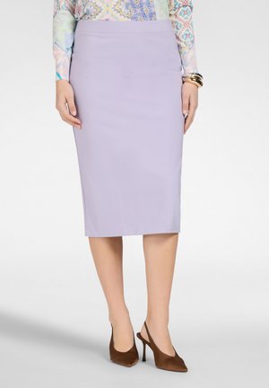 Kvinde iført en lyse lilla knælang pencilskørt, farverig mønstret bluse, guldarmbånd og brune høje hæle med åben hælkappe.