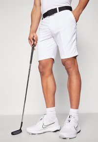 Bijele golf hlače, crni remen s logom, uparene s bijelim Nike golf cipelama i crnim golf palicom. Glatka tkanina, kroj koji ističe figuru i čiste linije.
