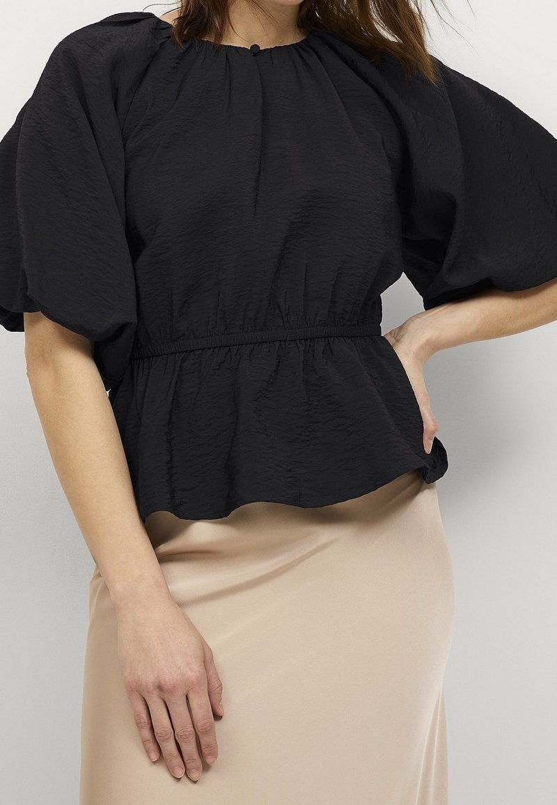 Black, crinkled fabric top with a round neckline, puffed sleeves, and elastic waistband, paired with a beige skirt.
