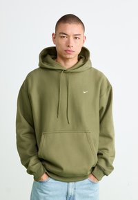 Ung mand med kort hår iført en olivengrøn Nike hoodie og lyseblå jeans, stående med hænderne i lommerne mod en hvid baggrund.