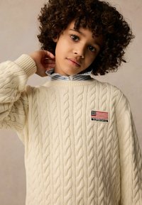 Cremefarbener Zopfmuster-Pullover mit rundem Ausschnitt, gerippten Bündchen und einem Aufnäher mit einer amerikanischen Flagge. Über einem hellblauen gestreiften Hemd getragen.