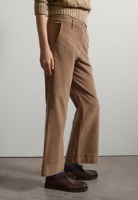 Pantalones de pana marrón de pernera ancha con dos bolsillos frontales, un cierre de botón y una tela texturizada. Combinados con zapatos oscuros y calcetines coloridos.