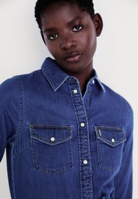 Denimskjorte i mørk blå med knappekrage, to brystlommer og hvite trykknapper. Har kontrasterende søm for detalj.
