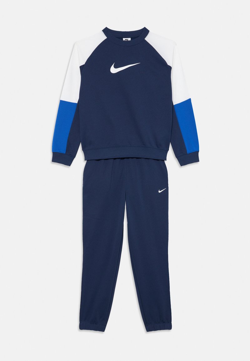 Survêtement bleu marine avec des manches colorées en blanc et bleu, présentant des logos Nike en blanc sur la poitrine et la cuisse gauche.