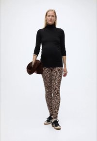 Έγκυος γυναίκα φορώντας μαύρο ζιβάγκο, κολάν με λεοπάρ σχέδιο και αθλητικά παπούτσια, κρατώντας ένα καφέ σακάκι σε απλό λευκό φόντο.