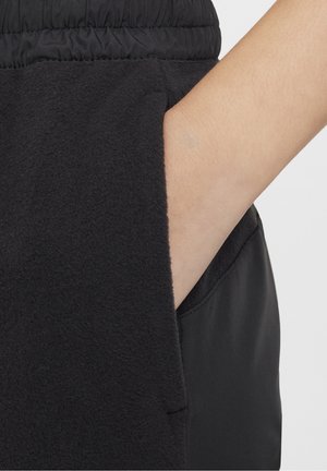 Hånd placeret inde i sidelommen på sorte fleecebukser med elastisk linning, der viser stoftekstur og syningsdetaljer.