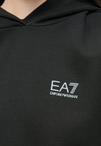 Czarna bluza z kapturem, miękką teksturą materiału i białym logo EA7 Emporio Armani na piersi. Prosty i nowoczesny design.