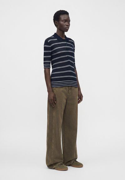 Personne debout portant un polo rayé bleu marine et blanc, un pantalon marron à jambes larges et des baskets marron sur un fond blanc uni.