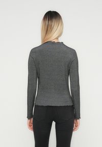 Donna con lunghi capelli biondi, che indossa un top aderente a maniche lunghe grigio scuro a coste e jeans neri, in piedi di spalle contro uno sfondo bianco.