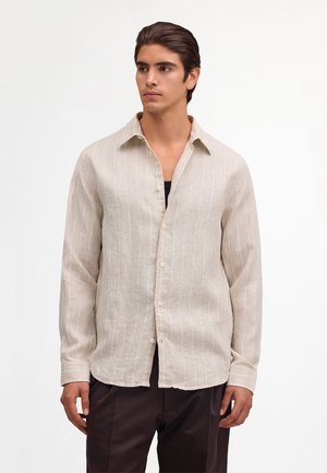 Ung mand iført en beige, langærmet, knappet skjorte med tynde lodrette striber over en sort indertrøje og mørke bukser.