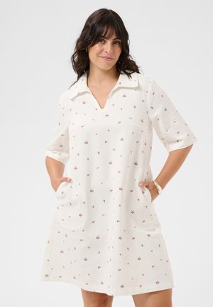 Vrouw met donker haar die een witte jurk tot op de knie draagt met kleine rode bloemenprint, halve mouwen, en handen in de zakken, licht glimlachend.