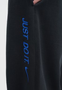 Melnas sporta bikses ar zilā izšuvuma tekstu "JUST DO IT." un Swoosh logotipu sānos, izgatavotas no mīkstas auduma ar gludu tekstūru.