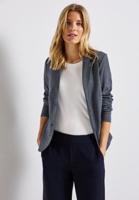 Šedý blazer s hladkou texturou, na jedno knoflíček a s ohrnutými rukávy, který je kombinován s bílou košilí a tmavě modrými kalhotami.