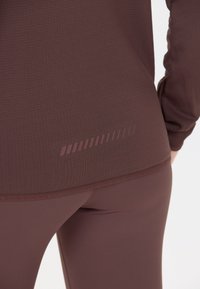 Maroonin värinen teksturoitu urheilu-toppi, jossa on kaareva helma ja huomaamaton raidallinen kuosi takana vyötärön kohdalla. Sujuva materiaali.