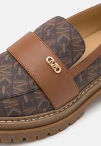 Mocassin marron et foncé à motif avec une semelle épaisse, une bride en cuir et un petit logo métallique doré sur le dessus.
