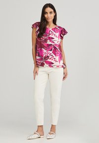 Blusa floreale con motivi rosa e bianchi, maniche corte a balze, abbinata a pantaloni slim di colore chiaro e scarpe slip-on decorate.