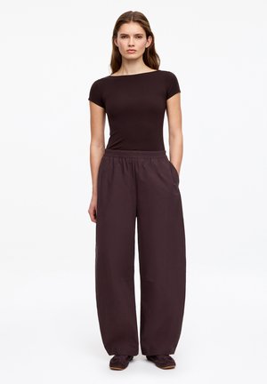 Kobieta stoi przodem, ubrana w dopasowaną czarną bluzkę z krótkim rękawem i luźne ciemne szerokie spodnie, trzymając ręce w kieszeniach.