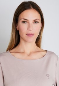 Jasnoróżowa koszula z długim rękawem, z zaokrąglonym dekoltem i subtelnym logo na piersi, wykonana z miękkiego, gładkiego materiału.